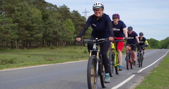 Four people are cycling along a quiet country road. All are wearing Cycling UK-branded T-shirts