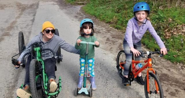 A woman on a recumbent trike is riding with two young girls, one on a scooter, the other on a bike