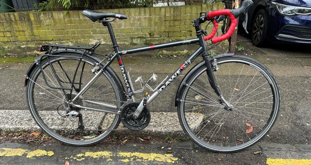 A black Dawes Galaxy touring bike with red bar tape is propped against a curb with the front wheel in a pothole