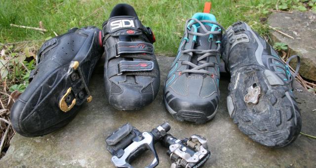 Four different types of clipless cycling shoes, with one lying on its side to show the cleat. In front are two types of clipless pedals