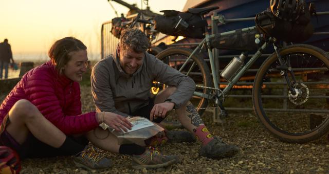 Two people study a map on a pebbly beach with bike propped up behind them