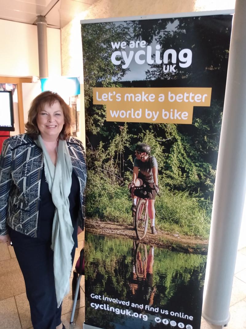 A woman wearing a patterned jacket, blue trousers and pale blue scarf is posing next to a Cycling UK poster