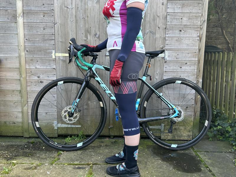 A woman in cycling jersey and shorts with Castelli arm and knee warmers is standing in front of a Felt road bike leaning against a garden shed