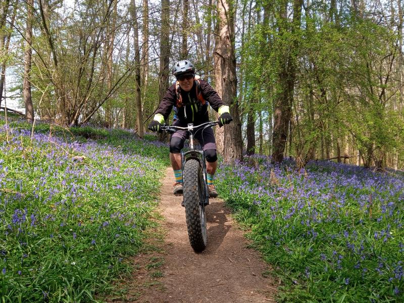 A man is riding a fat bike on a trail through a bluebell wood. He's wearing mountain biking gear, sunglasses and a helmet and is smiling at the camera