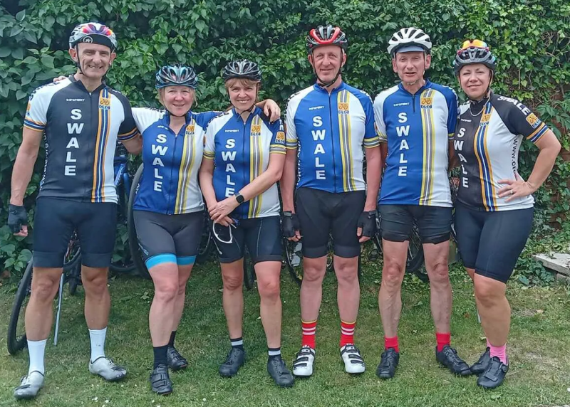 A group of people are standing on grass with a hedge behind them. They are all wearing CTC Swale cycling kit, which is blue, white and gold. They all have helmets on.