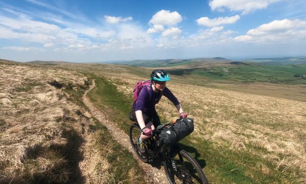 A woman is cycling a loaded mountain bike on a bridleway on a high ridge. She is wearing mountain bike gear and a helmet. It's a sunny day.