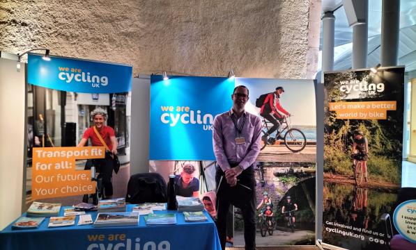 A man in shirt and trousers with a name tag round his neck and holding a clipboard. He is at an exhibition stand with Cycling UK branded posters and table